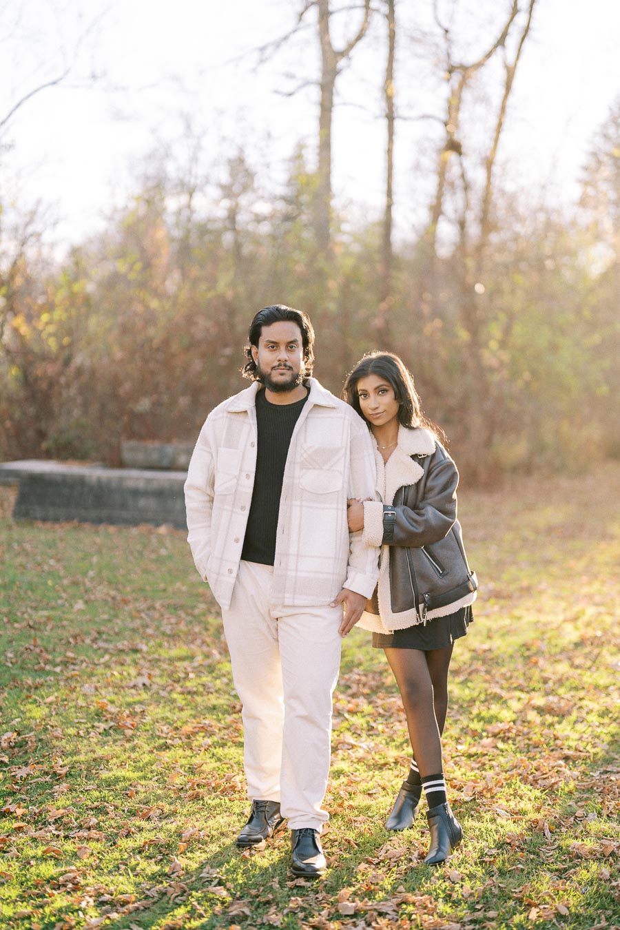 A couple standing together in a sunlit autumn park, both wearing stylish jackets, surrounded by fallen leaves and bare trees in the background.