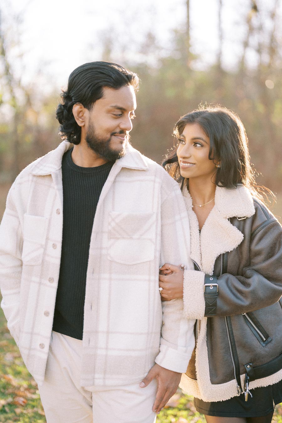 A couple smiling and embracing, dressed in cozy winter jackets, enjoying a sunny day in a park.