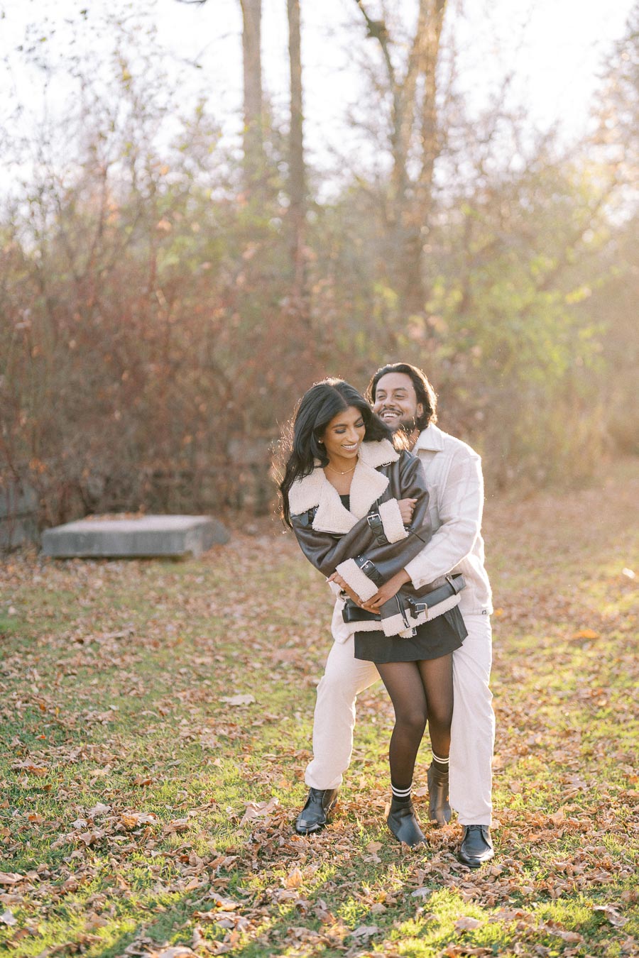 A couple enjoying a playful embrace in an autumn forest setting, surrounded by fallen leaves and warm sunlight.