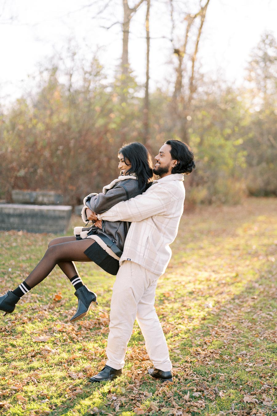 A couple enjoying a playful moment outdoors in autumn, with fallen leaves scattered on the grass and trees in the background, under a clear sky.