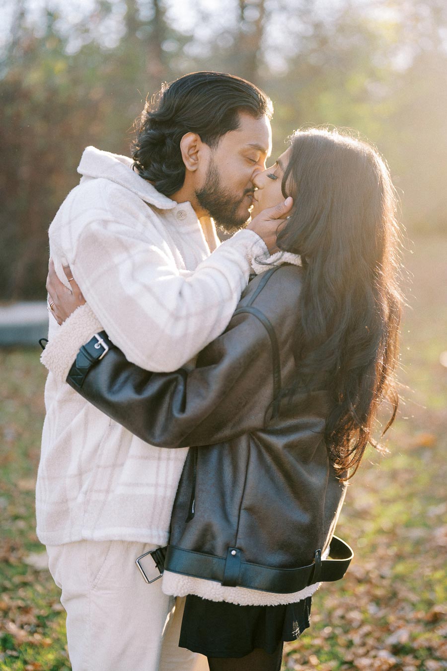 A couple softly kissing outdoors on a sunny day, surrounded by a natural, leafy background, wearing cozy jackets.