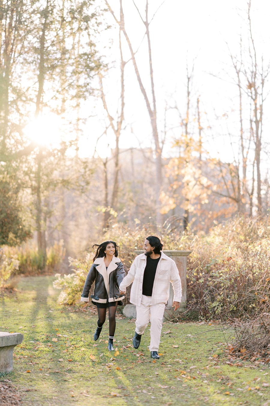 A couple walks hand in hand through a park with autumn foliage, bathed in warm sunlight. They wear stylish fall jackets, enjoying the serene outdoor setting.