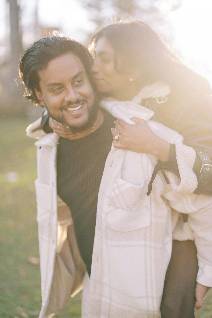 A couple enjoying a playful moment outdoors, with a woman kissing a smiling man's cheek while giving him a piggyback ride in a sunlit park.