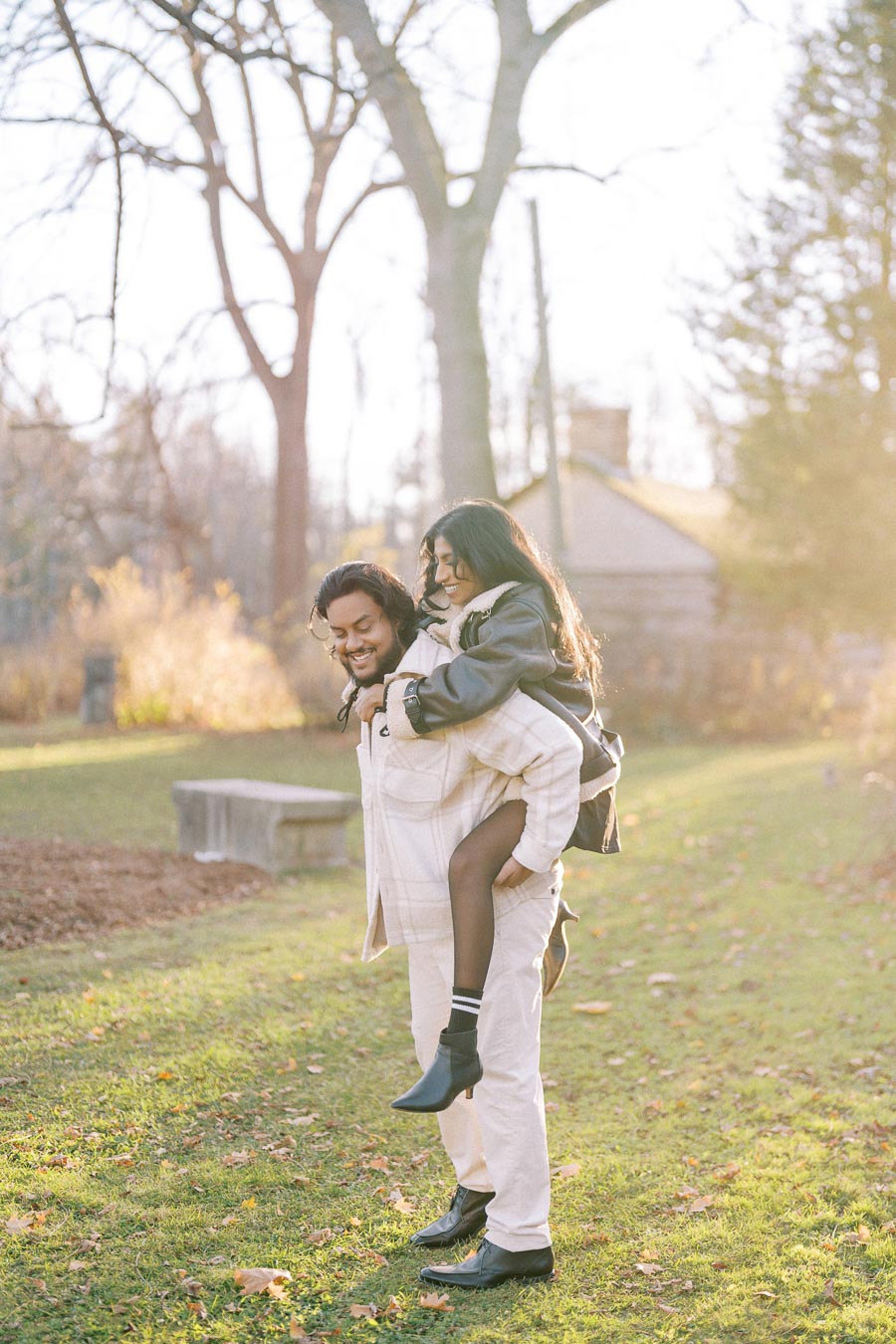 A couple enjoying a playful piggyback ride in a sunny park during autumn, surrounded by trees and fallen leaves.