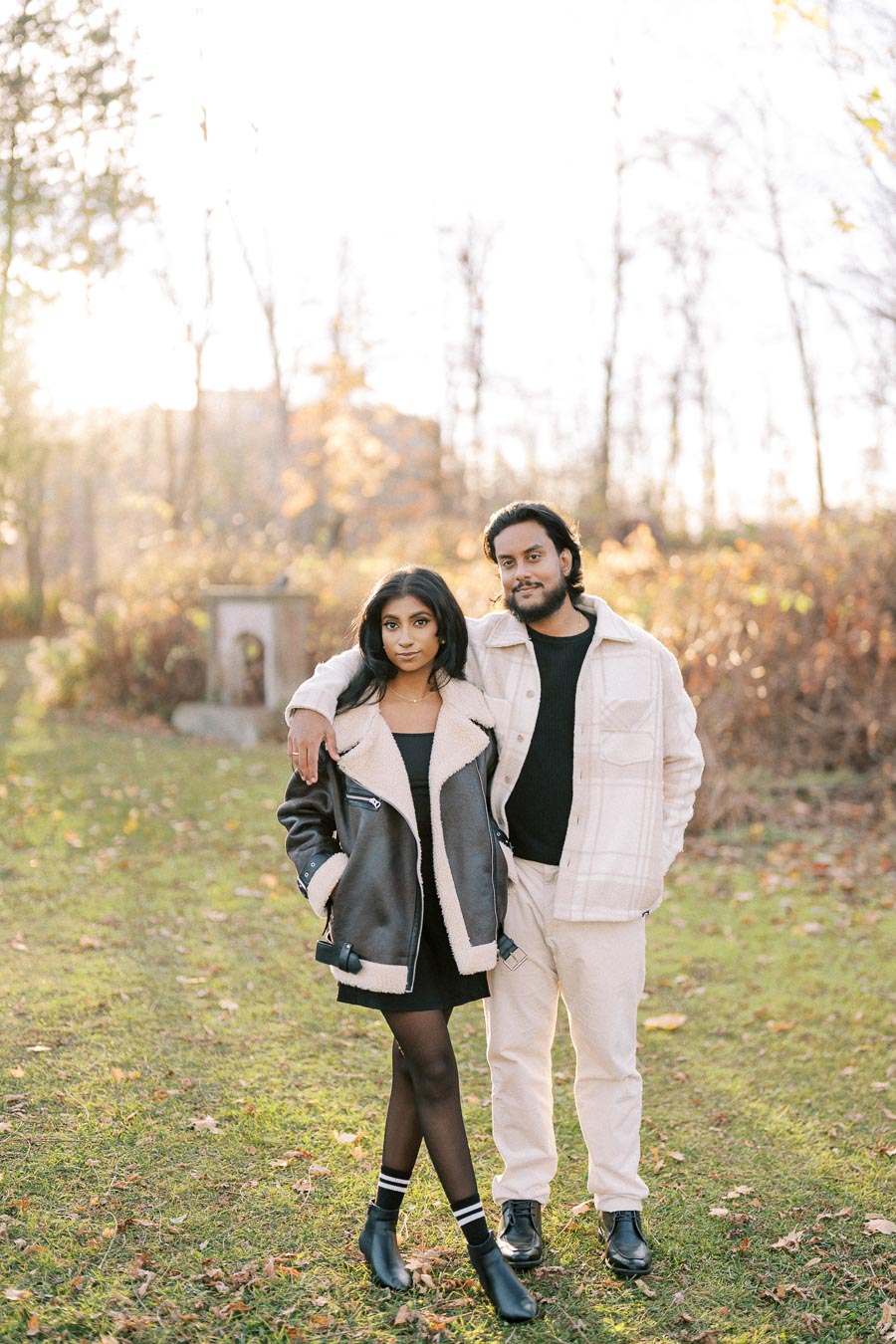 A couple standing in a sunlit autumn park, wearing stylish jackets and looking confidently at the camera.