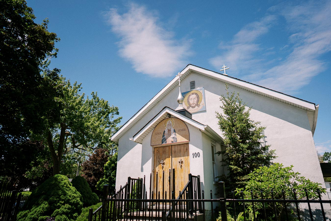 White church building with religious mosaics and a cross on the roof, surrounded by greenery and a sunny blue sky.