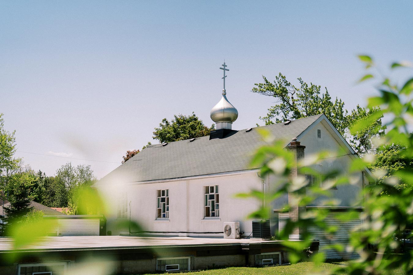 A small white church with a traditional Russian-style dome and cross seen through green foliage on a sunny day, showcasing a peaceful suburban setting.
