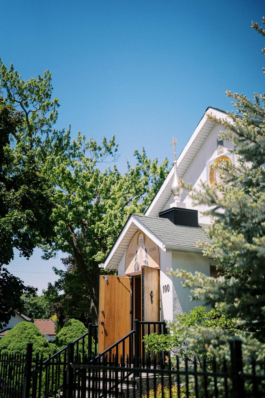 White church building with a cross-topped roof, open wooden doors, and surrounding green trees under a clear blue sky.