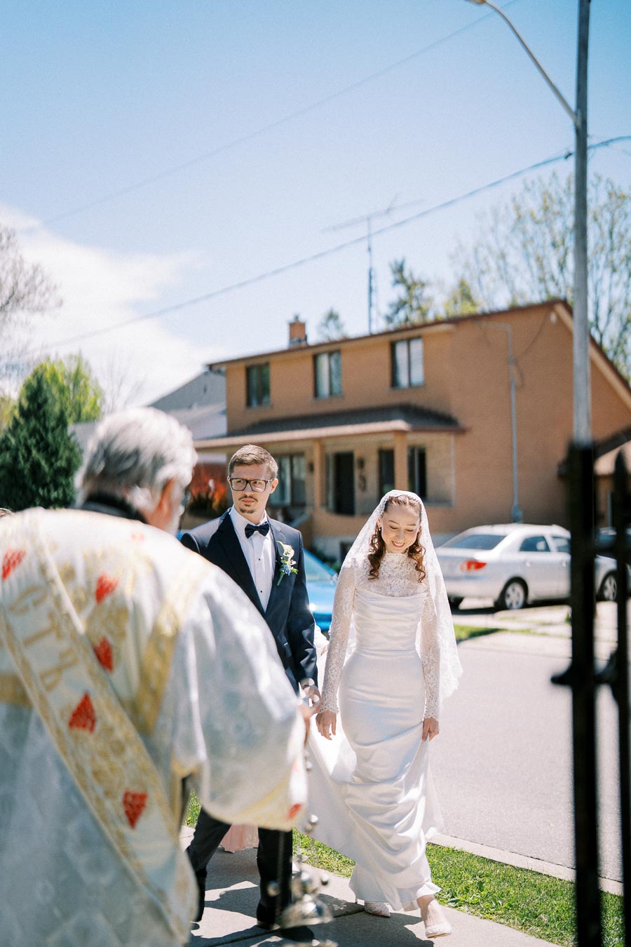 Wedding couple walks together outside after ceremony, with a clergy member leading them. The bride is in a white dress and veil, and the groom is in a black tuxedo. They are smiling, and the background shows suburban houses and a clear sky.