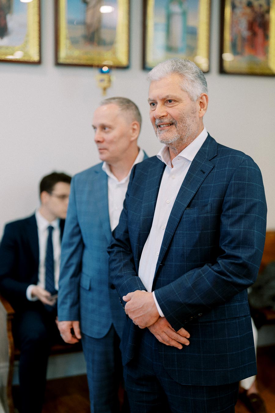 A group of well-dressed men in blue suits attending a formal event indoors, with framed artwork on the wall in the background.