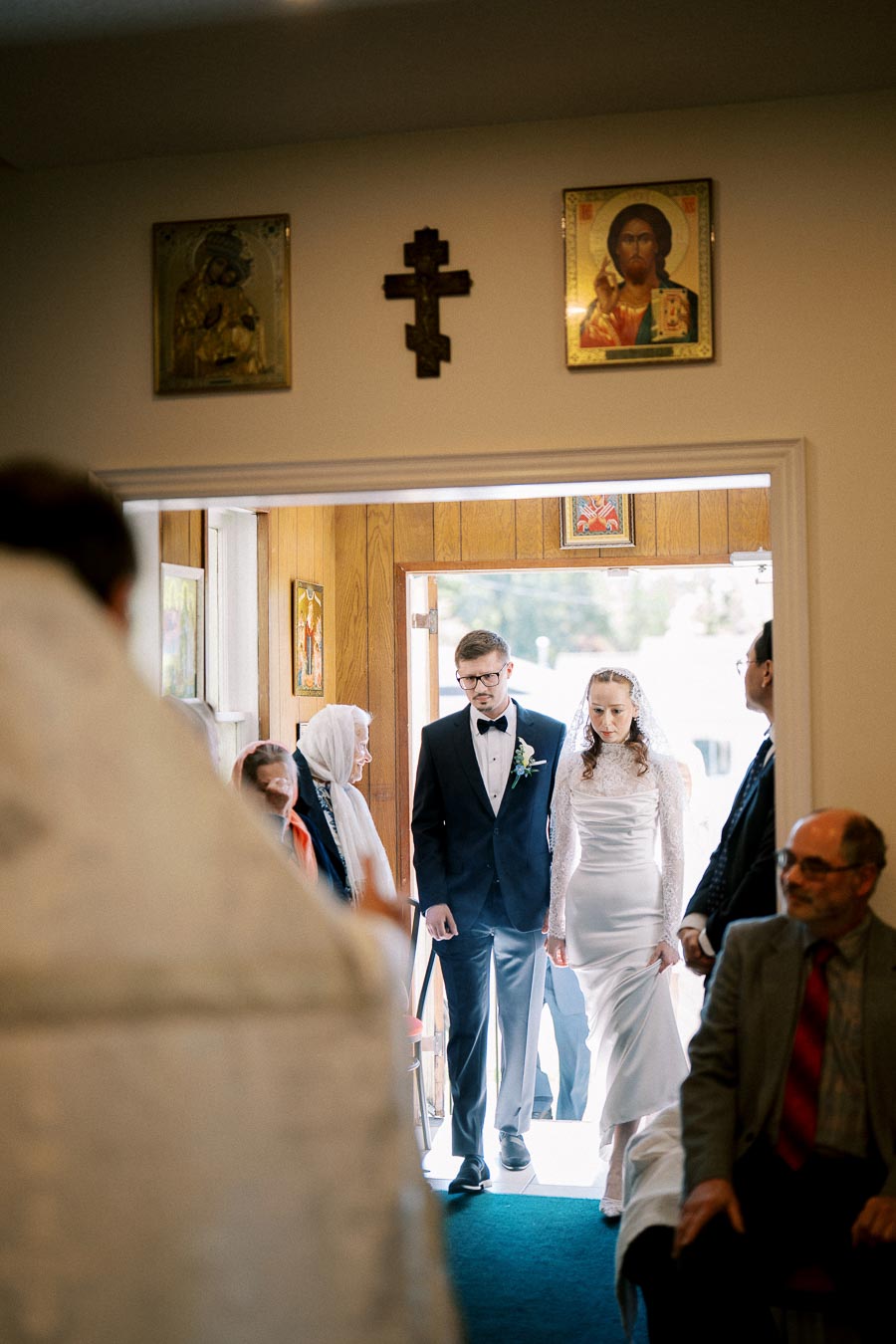 A bride and groom in formal attire entering a church for a wedding ceremony, with religious icons on the wall and guests in the background.