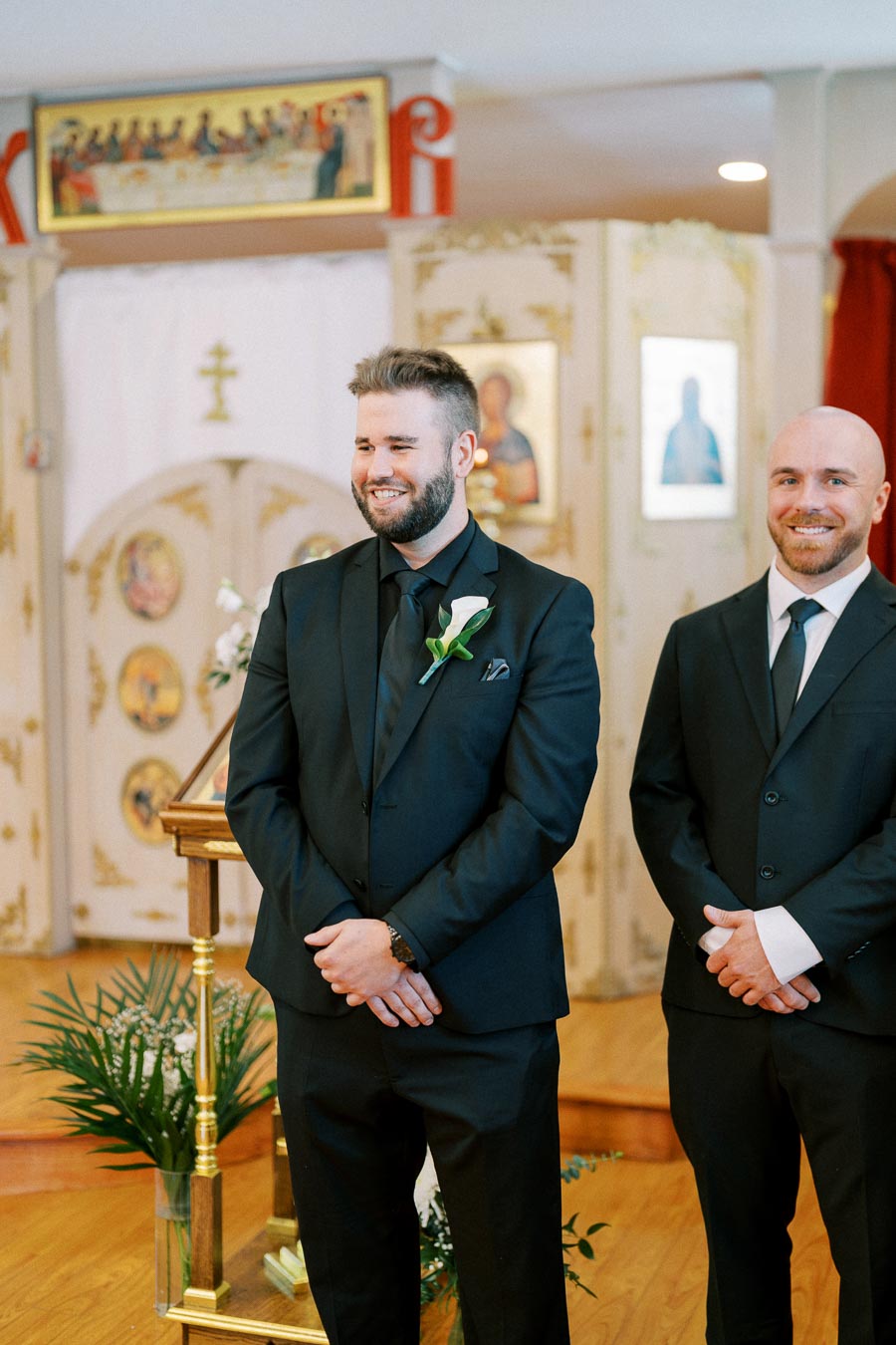 Two groomsmen in black suits, smiling during a church wedding ceremony, with ornamental backgrounds and religious iconography.
