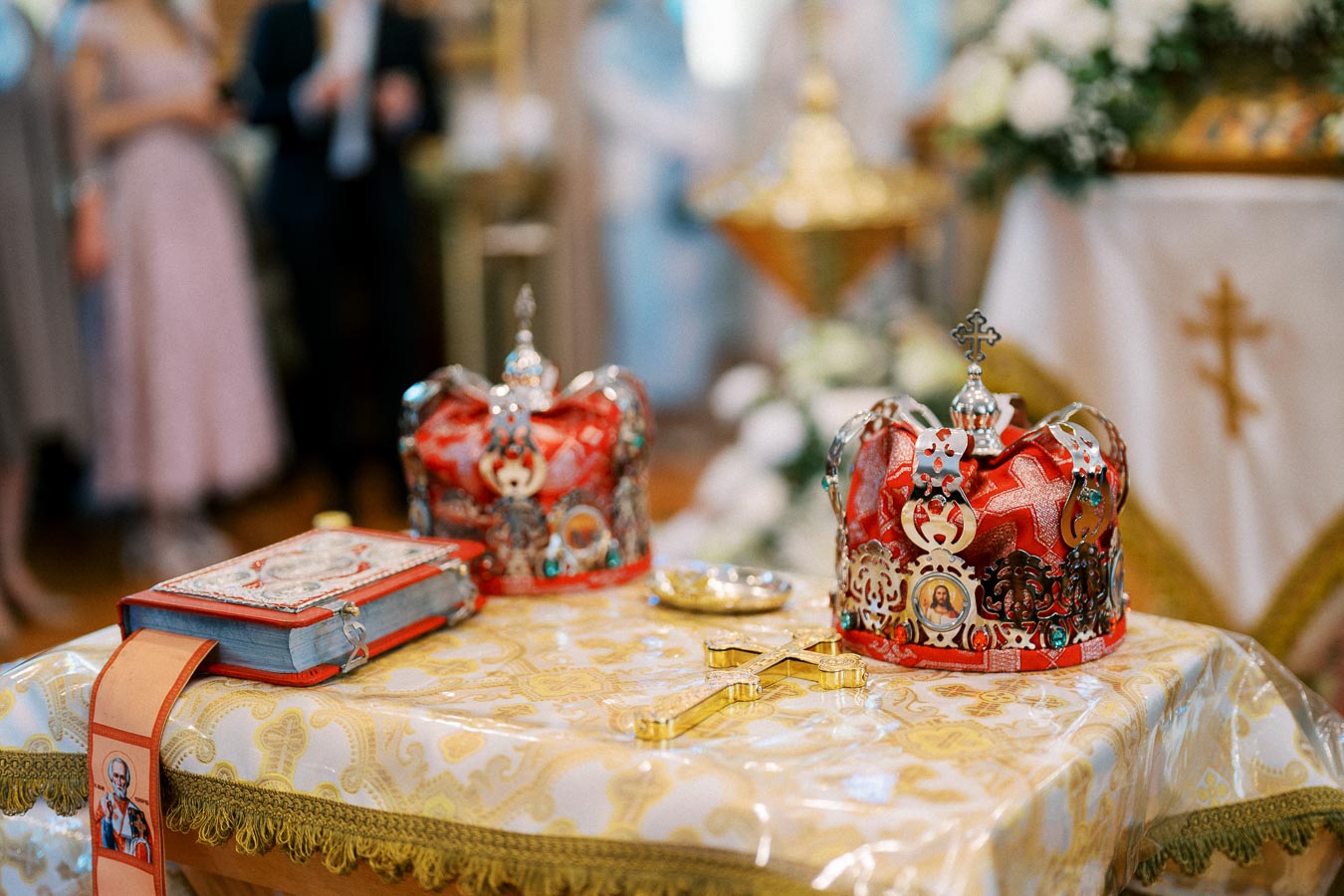 A close-up of ornate crowns and a decorative cross placed on a table with an embroidered cloth, set in a church environment.