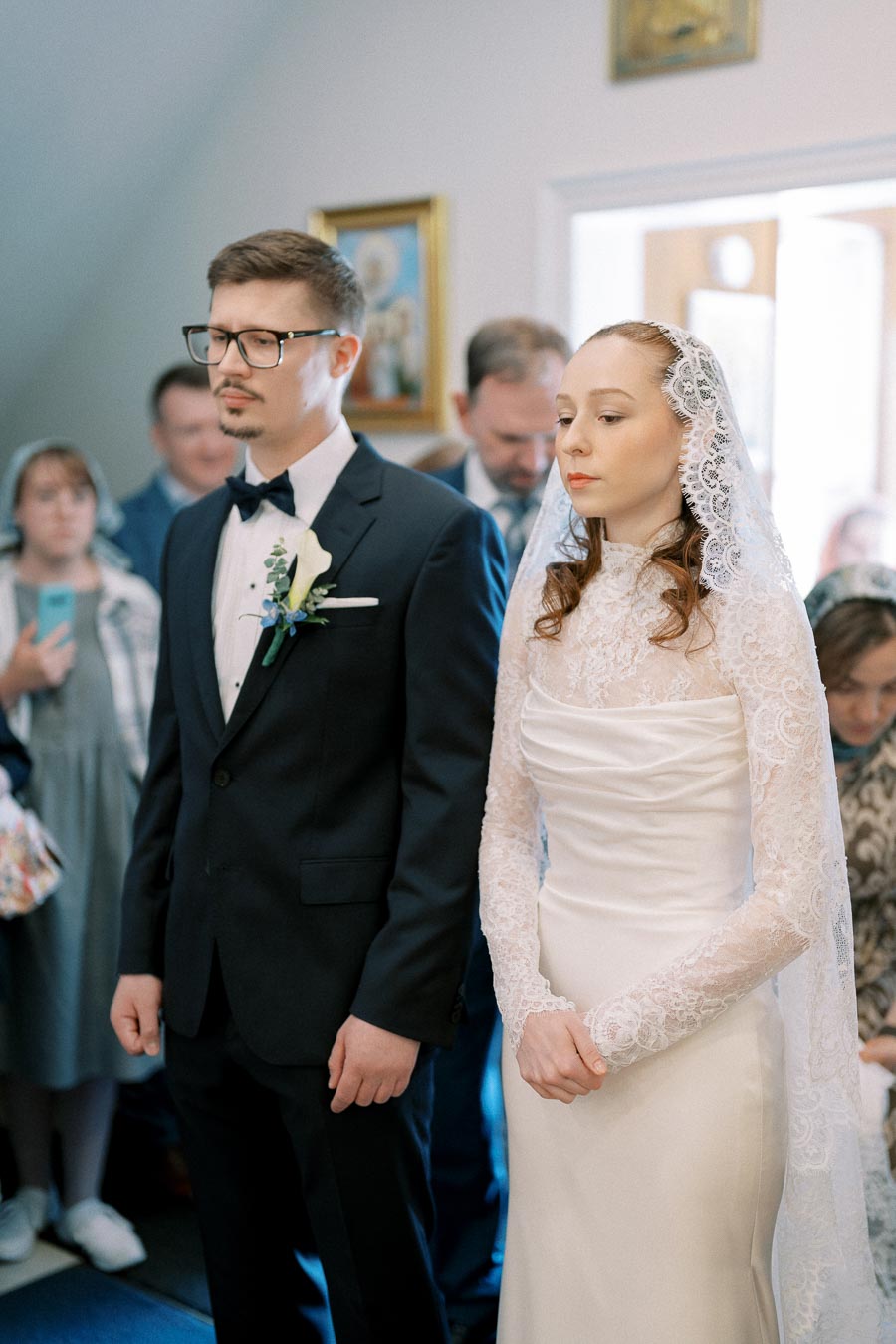 Couple standing during traditional wedding ceremony, bride in lace dress and groom in dark suit with boutonniere, with guests in background.