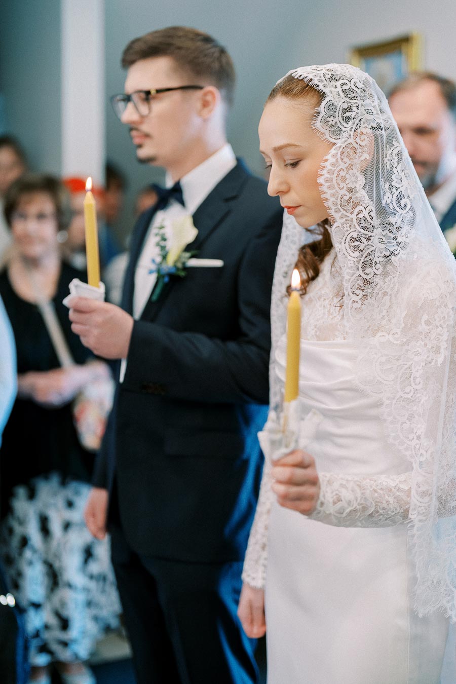 A bride and groom holding candles during a traditional wedding ceremony, with the bride wearing a lace veil.