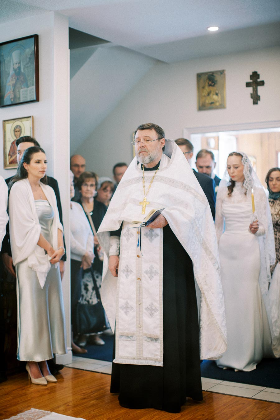 A religious ceremony inside a church, featuring a priest in traditional Orthodox attire holding a book, surrounded by a group of people. The setting includes religious icons on the wall and attendees in formal clothing, suggesting a special occasion or service.