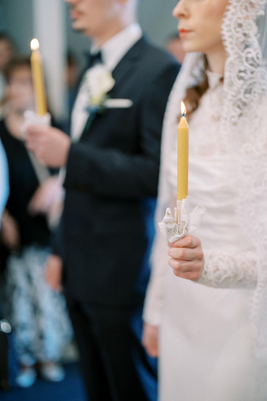 A bride and groom holding lit candles during a wedding ceremony, the bride wearing a lace veil and dress, and the groom in a suit with a boutonniere, surrounded by blurred guests in soft focus.