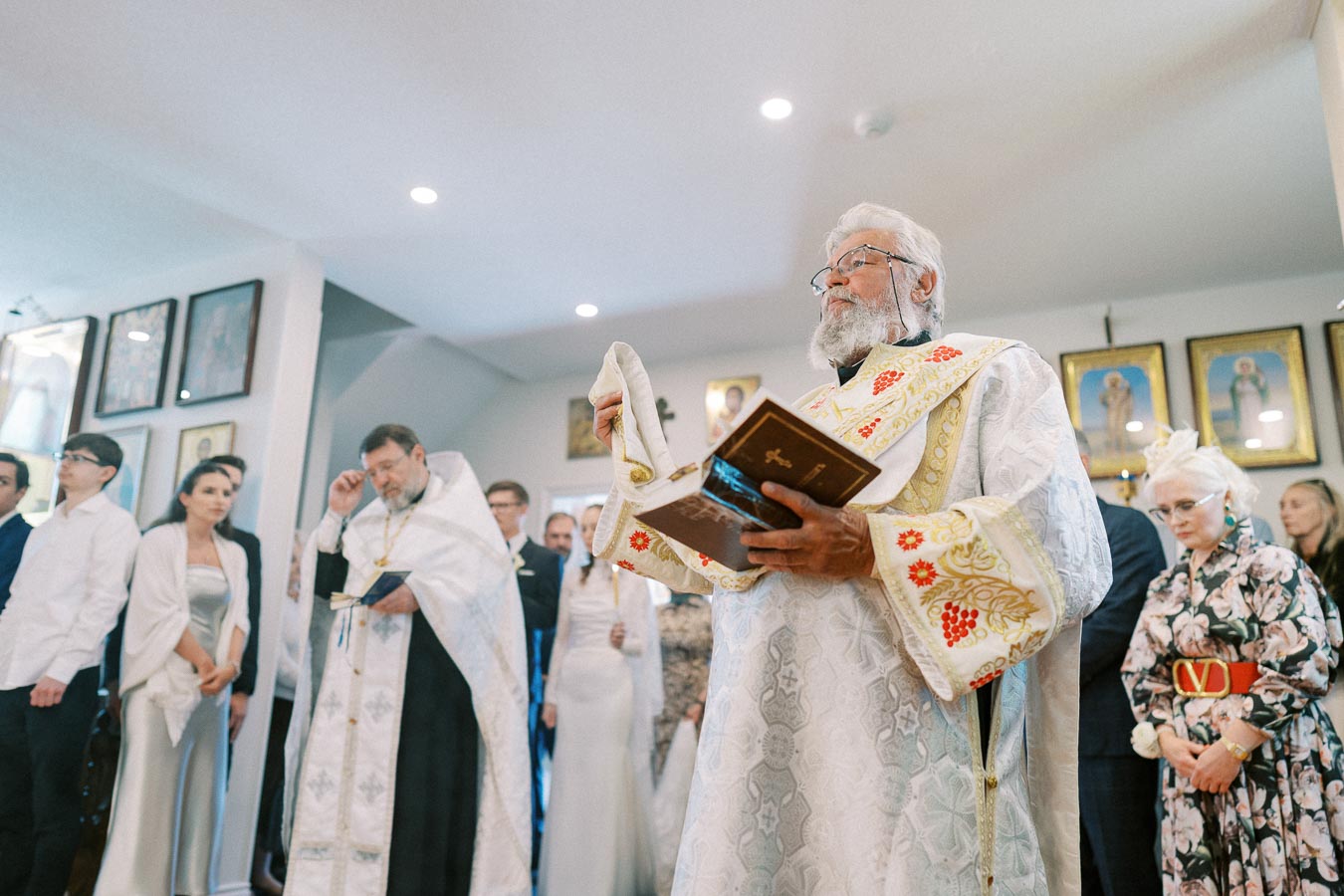 Elderly priest conducting religious ceremony in ornate vestments within a church, holding a sacred book, surrounded by congregants dressed formally, with religious icons on the wall in the background.