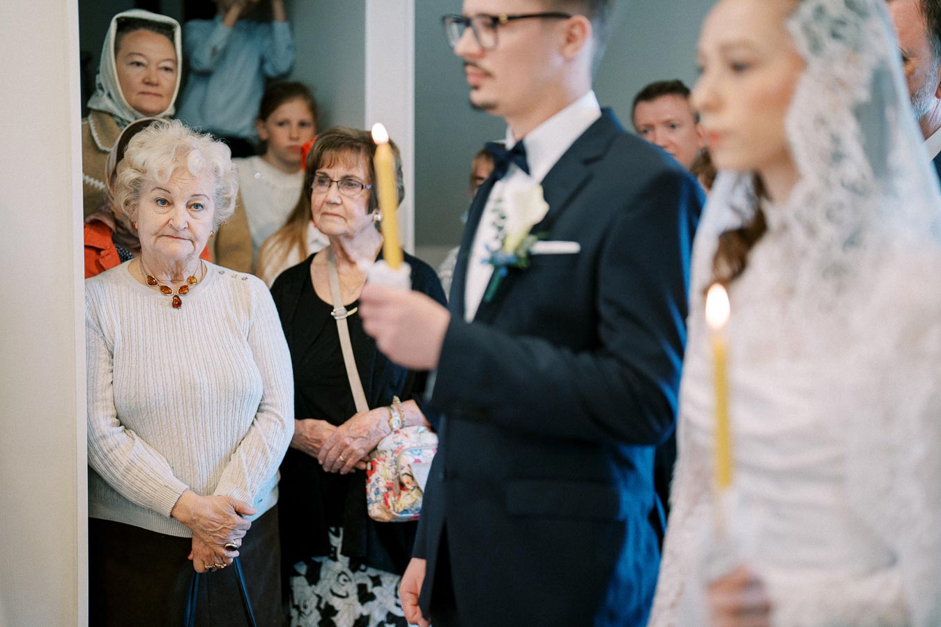 A group of people attending a formal event, with a man in a suit holding a candle and a woman in a white lace veil. In the background, older women and children watch attentively, suggesting a ceremonial or religious gathering.