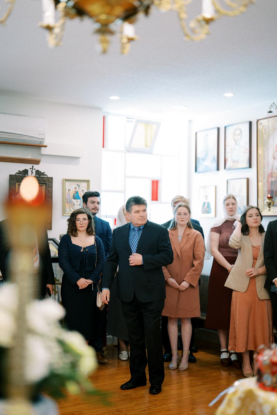 A group of people dressed in formal attire standing inside a room with framed portraits and religious icons on the wall, suggesting a ceremonial or religious gathering atmosphere.