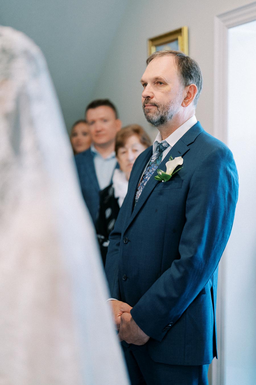 A man in a blue suit with a floral lapel pin stands solemnly, surrounded by a group of people, at a formal indoor event.