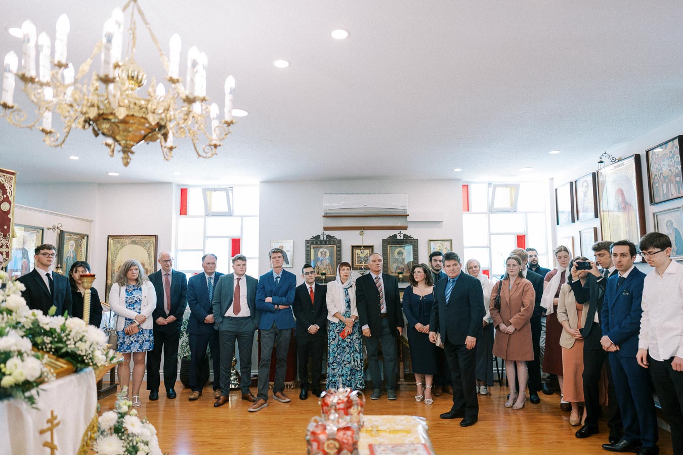 A diverse group of people in formal attire attending a religious ceremony in a well-decorated room with chandeliers and religious icons on the walls.