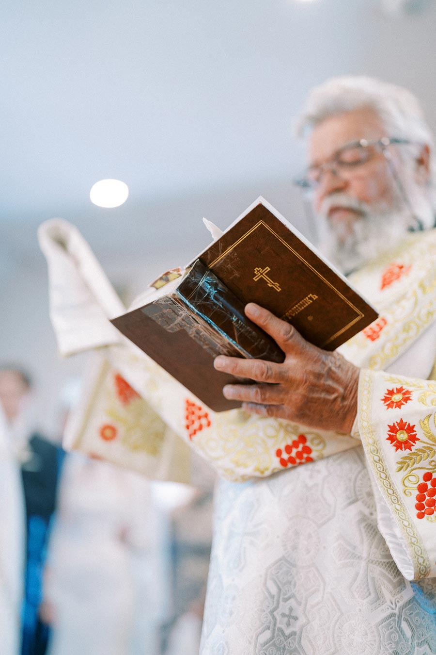 Elderly priest in ornate robe reading a religious book during a church ceremony.