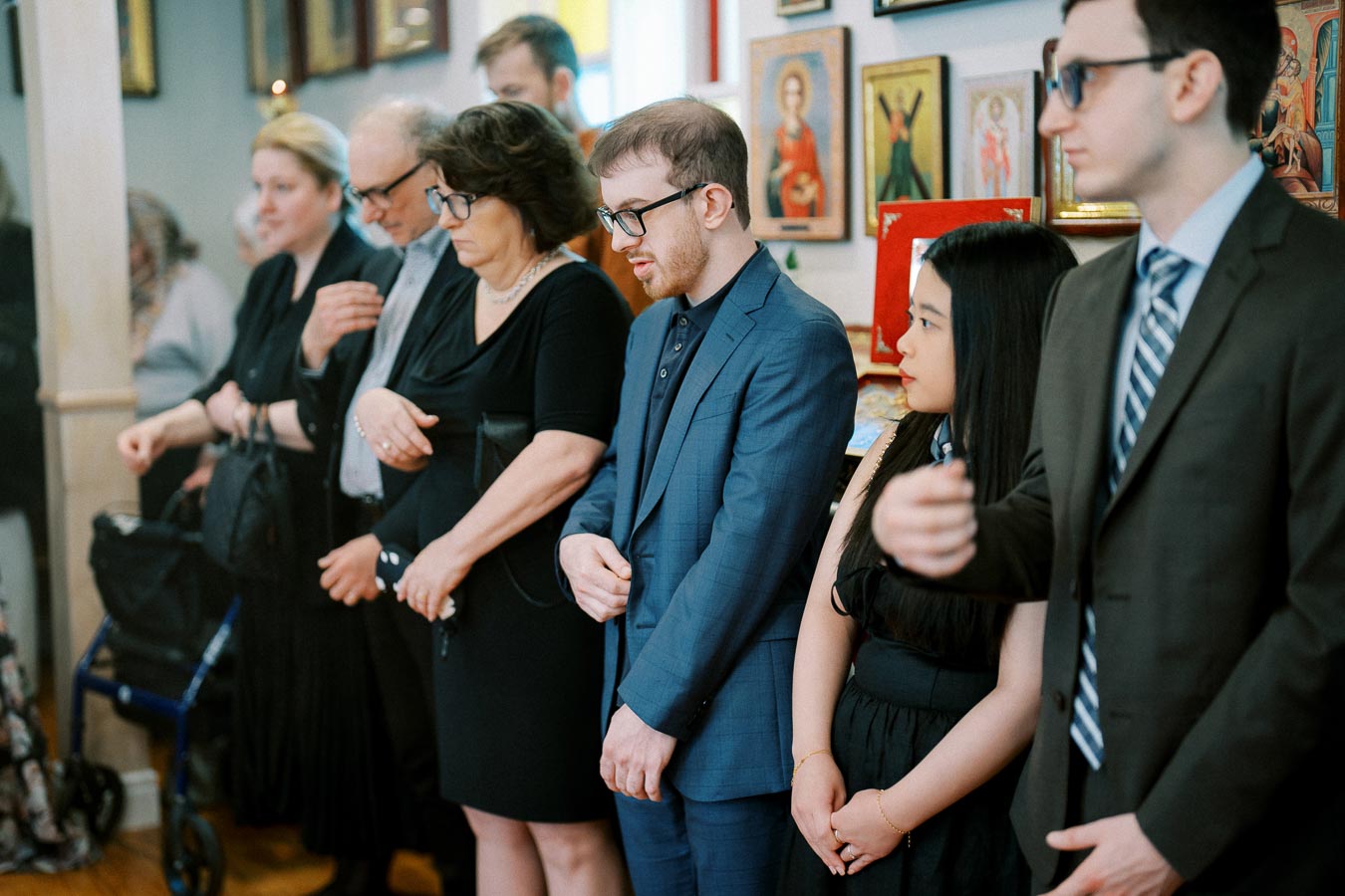 A group of people standing in a solemn setting, dressed in formal attire, possibly attending a ceremony or event, with art and religious iconography in the background.
