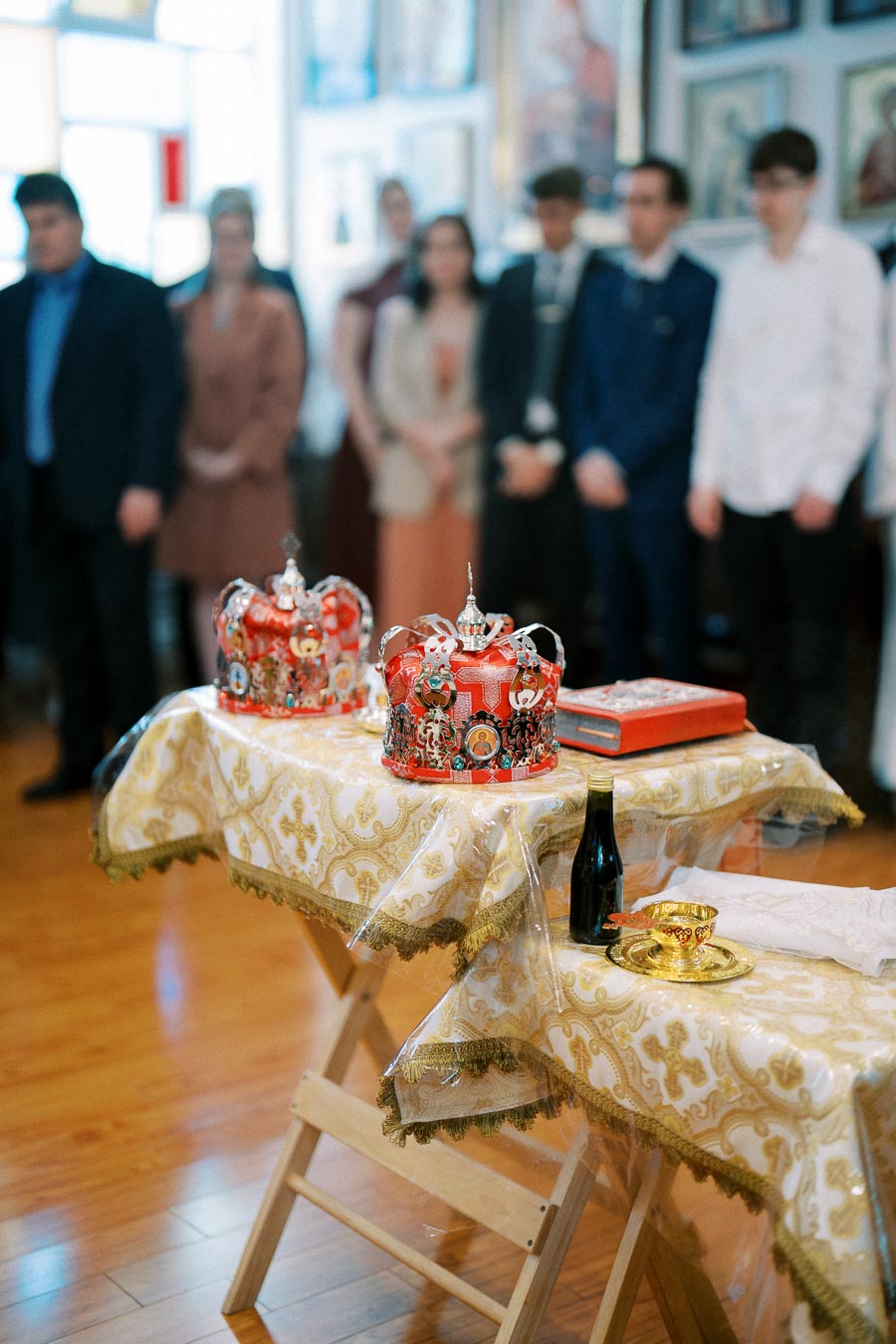 An ornate church wedding ceremony setup featuring two decorated crowns on a table with a gold-patterned cloth, surrounded by religious items, with attendees blurred in the background.