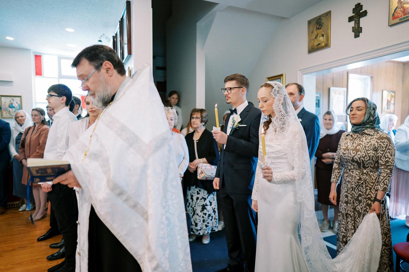 Orthodox wedding ceremony with a bride in a lace veil and groom holding candles, surrounded by guests and a priest reading from a book in a richly decorated church interior.