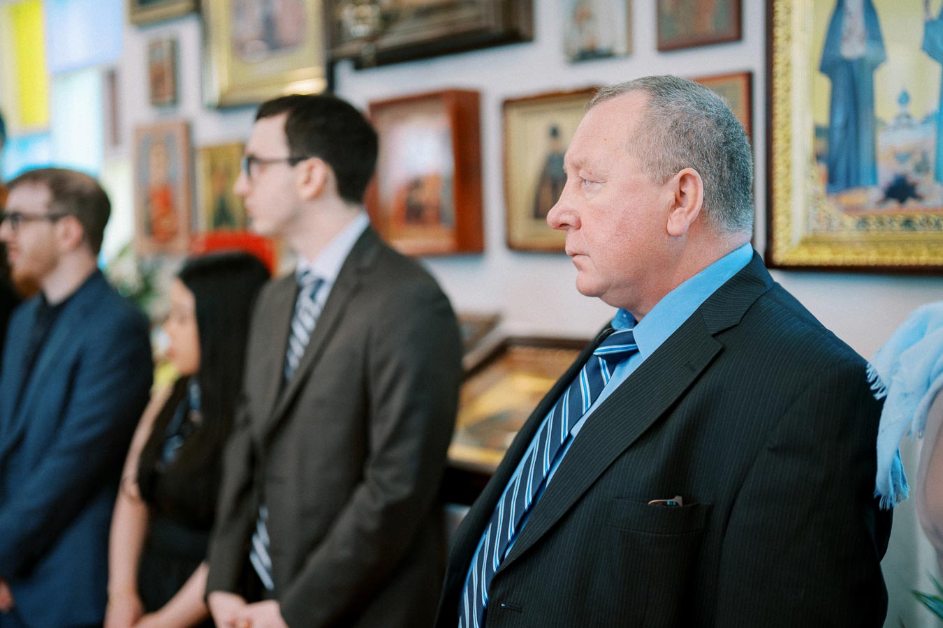 A group of people in formal attire standing in a room adorned with framed artwork and religious icons.