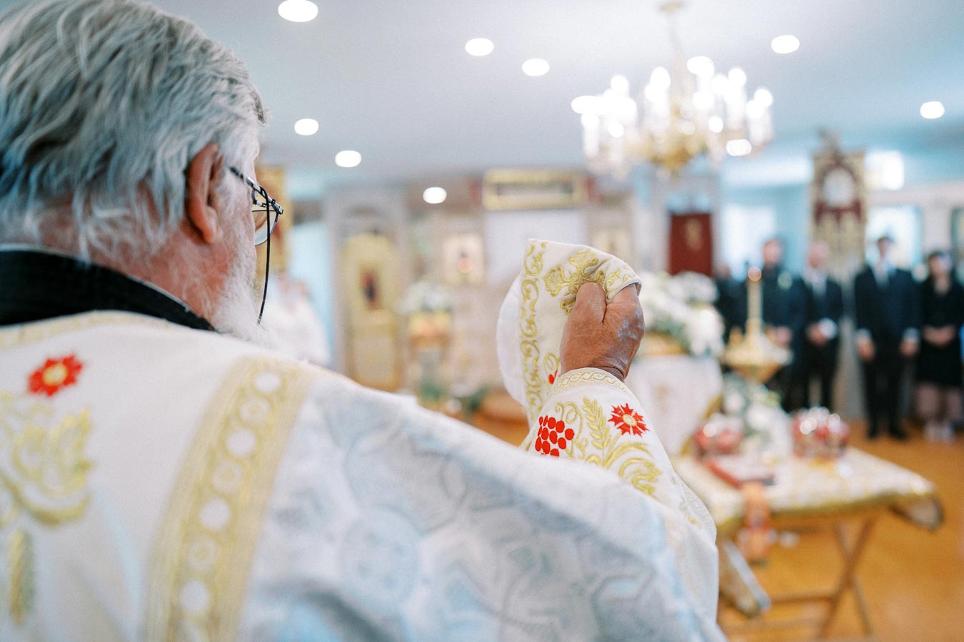 Elderly priest in ornate robe holding a decorated cloth during a religious ceremony inside a well-lit church with chandeliers and people in the background.