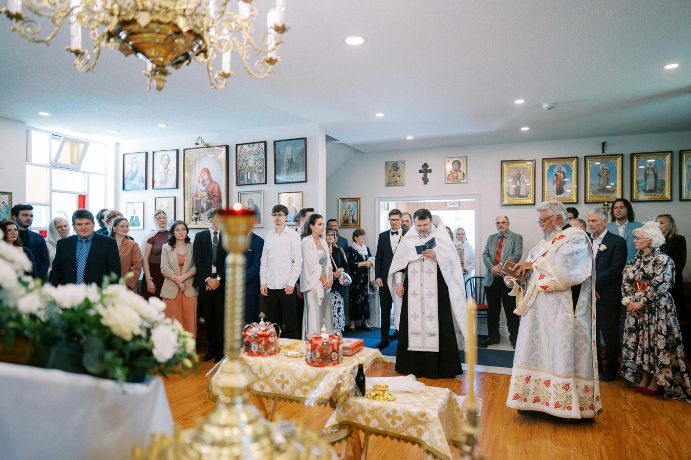 A religious ceremony inside a church with priests in traditional vestments conducting a service. Attendees, dressed in formal attire, line the walls adorned with religious icons and paintings. A chandelier and decorative table with ceremonial items are visible in the foreground, creating an atmosphere of reverence and tradition.