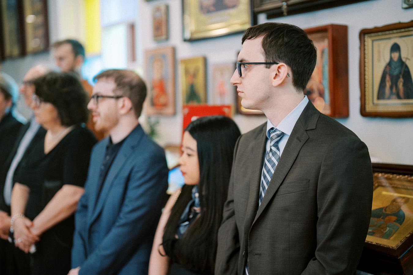 A group of people dressed formally stand attentively in an art gallery, surrounded by framed artwork and religious icons on the walls.