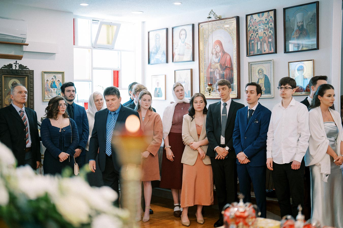 A group of people standing inside a room decorated with religious icons and paintings, dressed formally, appearing to participate in a ceremony, with blurred flowers and a candle flame in the foreground.