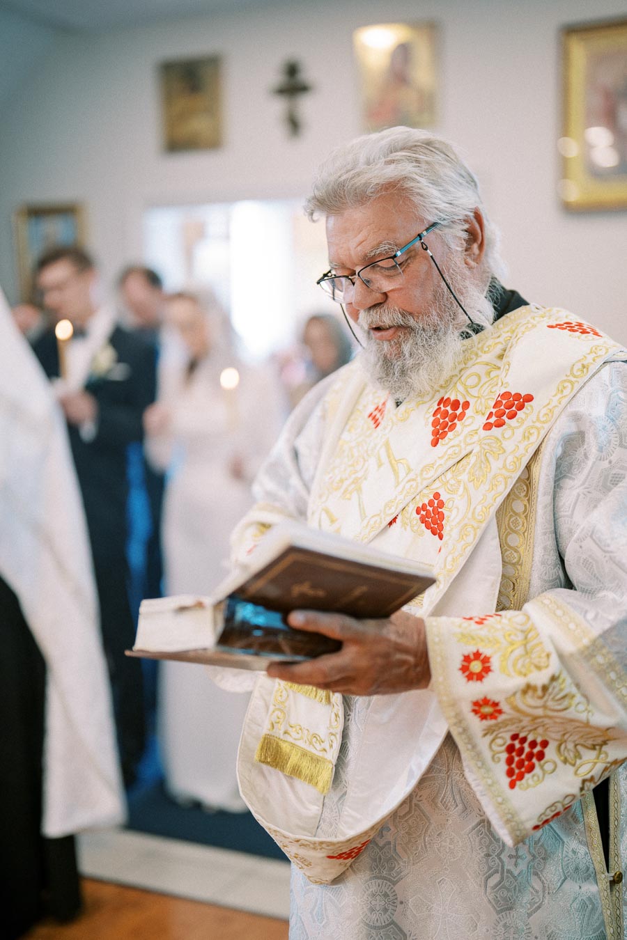 Elderly priest in ornate robe reads from a religious book during a wedding ceremony inside a church, with blurred guests holding candles in the background.