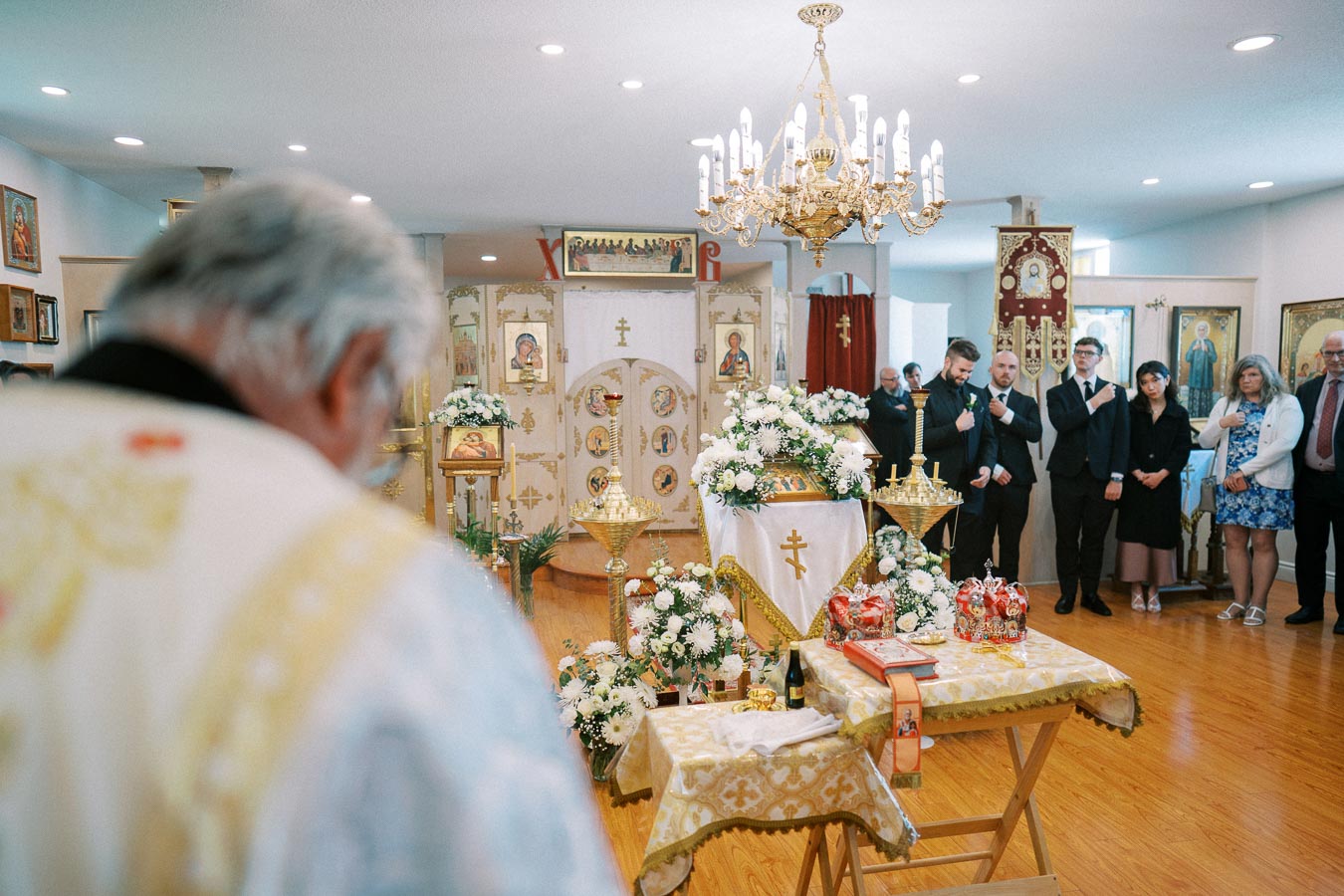Orthodox church service with priest and congregation, adorned altar with traditional icons and floral arrangements, warm lighting highlighting religious ceremony atmosphere.