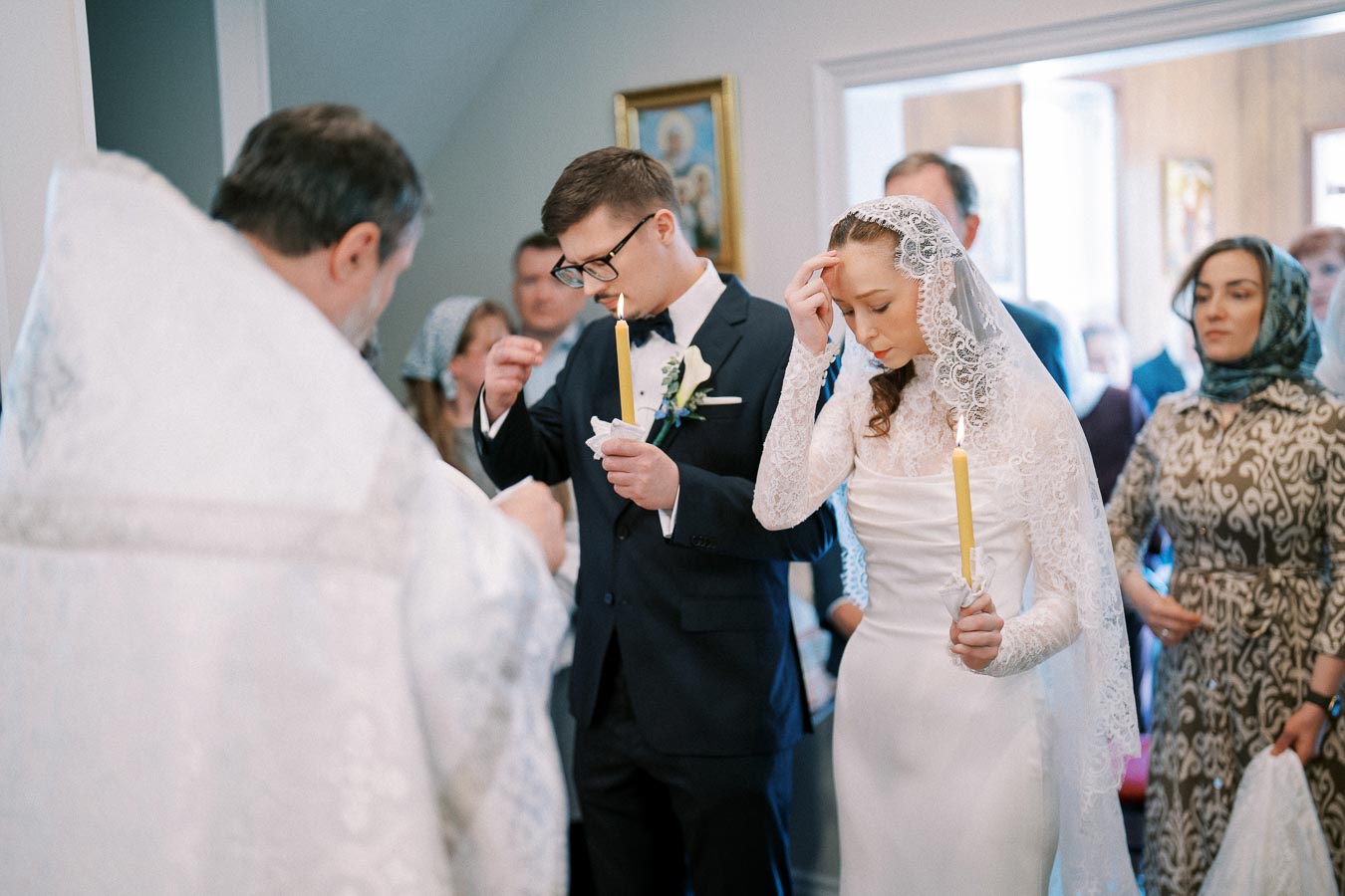 Bride and groom in traditional wedding attire holding candles during a religious ceremony in a church setting, surrounded by guests.