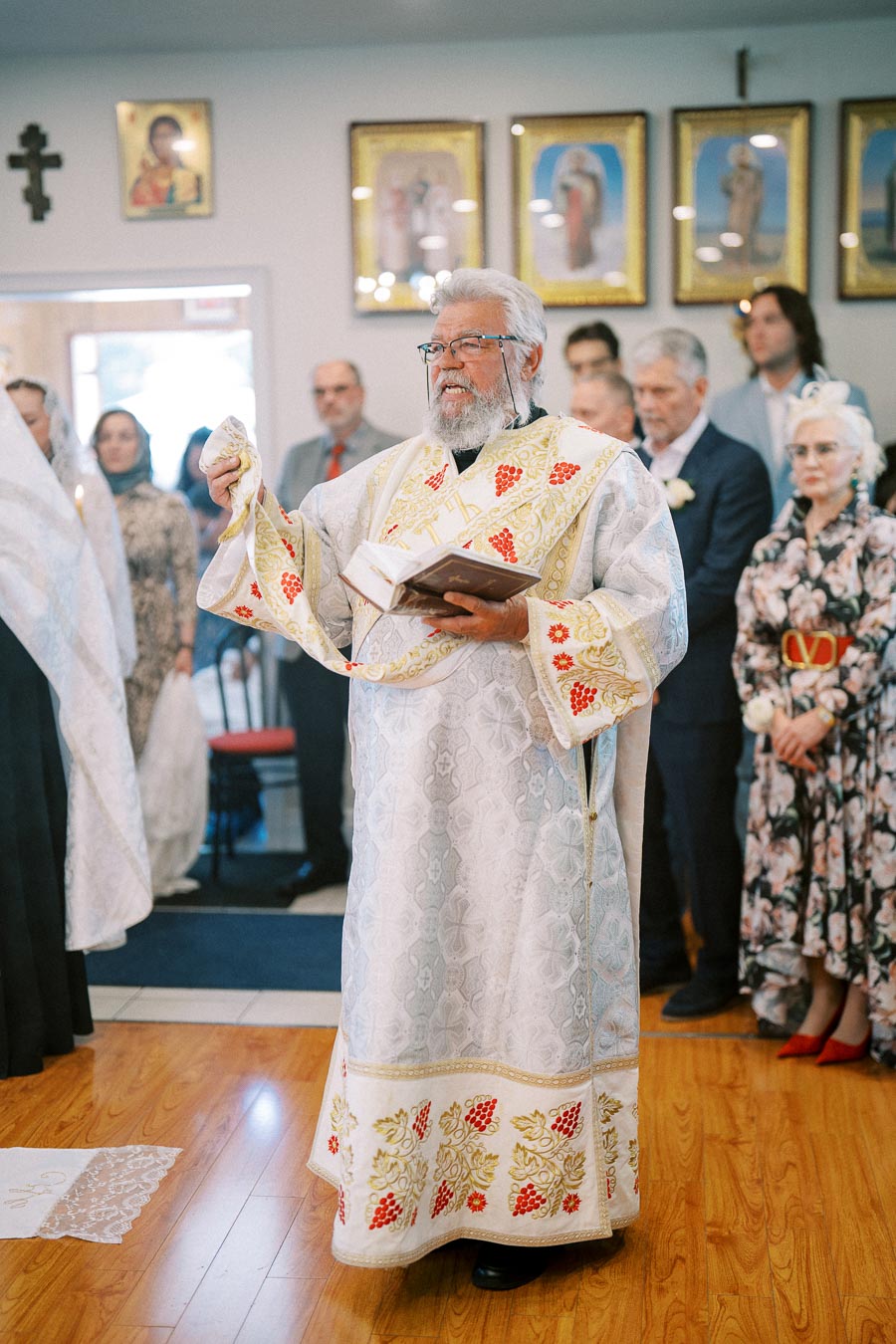 Priest conducting a religious ceremony in a church, wearing ornate vestments with red and gold embroidery, surrounded by attendees in formal attire.