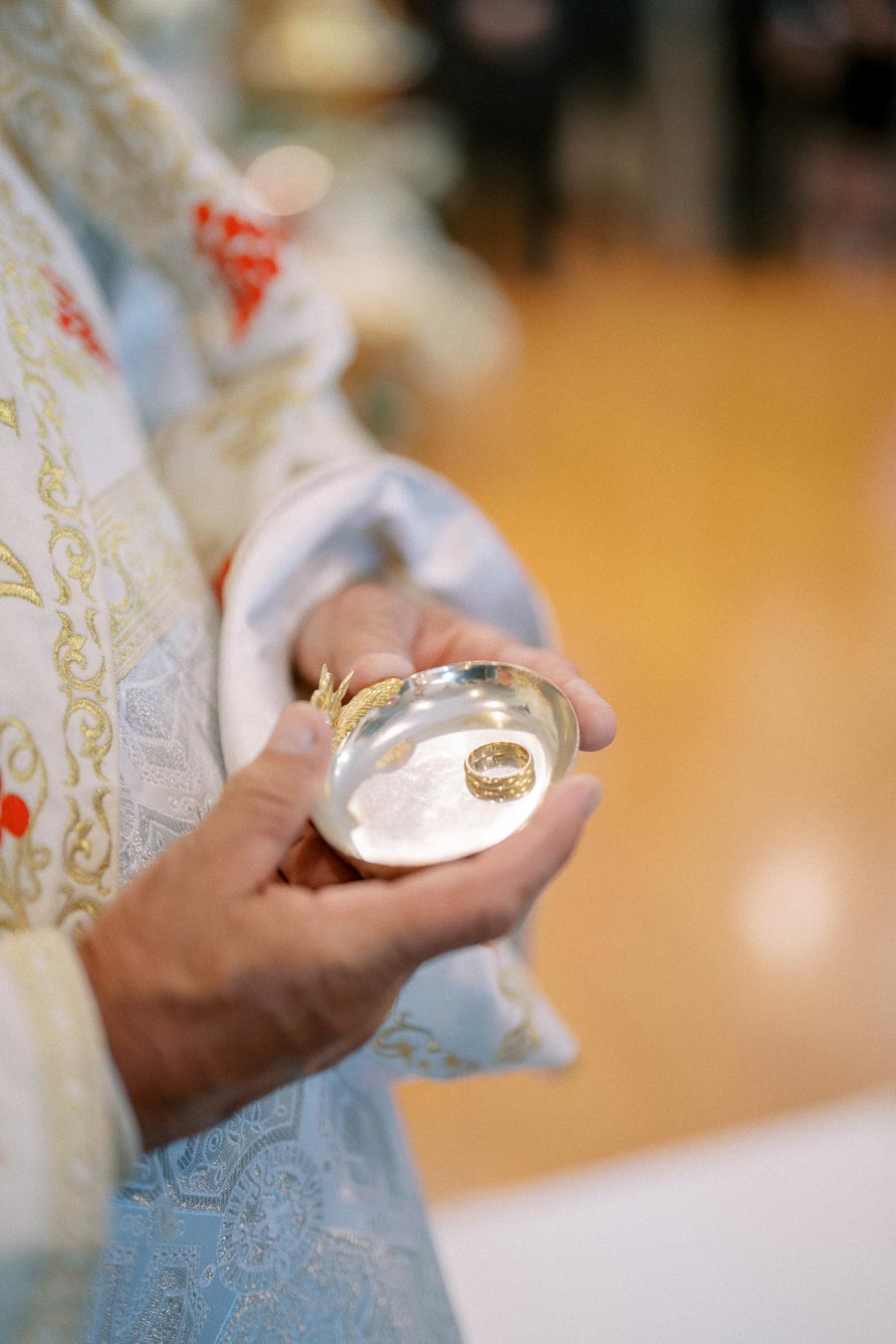 Close-up of a person wearing ornate ceremonial robes holding a silver dish with a gold wedding ring, symbolizing a traditional marriage ceremony.