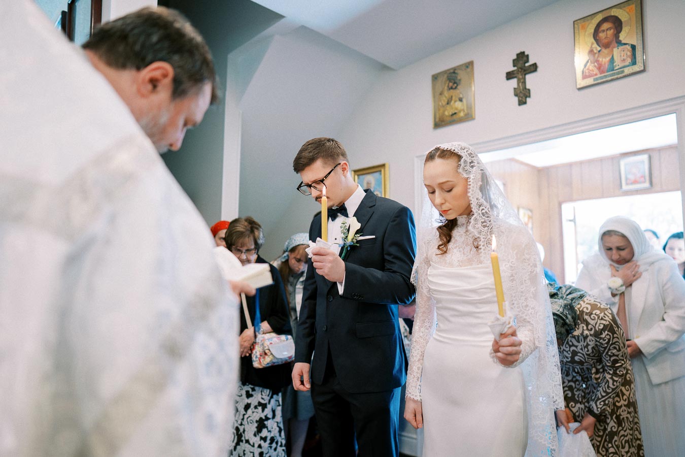 A bride and groom holding candles during an Orthodox wedding ceremony, surrounded by family and friends in a church adorned with religious icons.