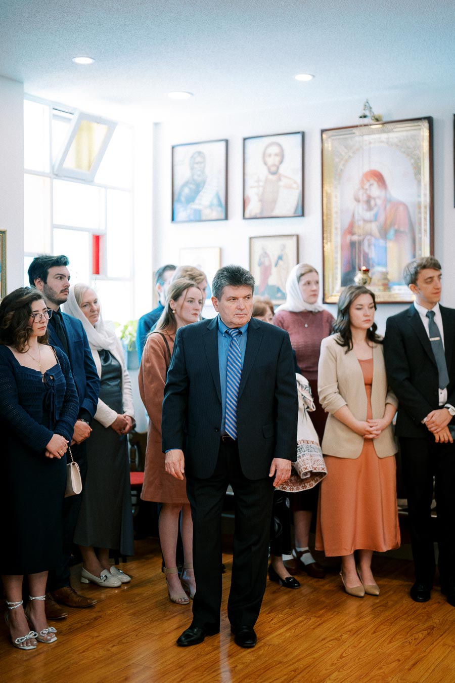 A group of people standing in a room with religious icons on the wall, dressed in formal attire, with a solemn expression.