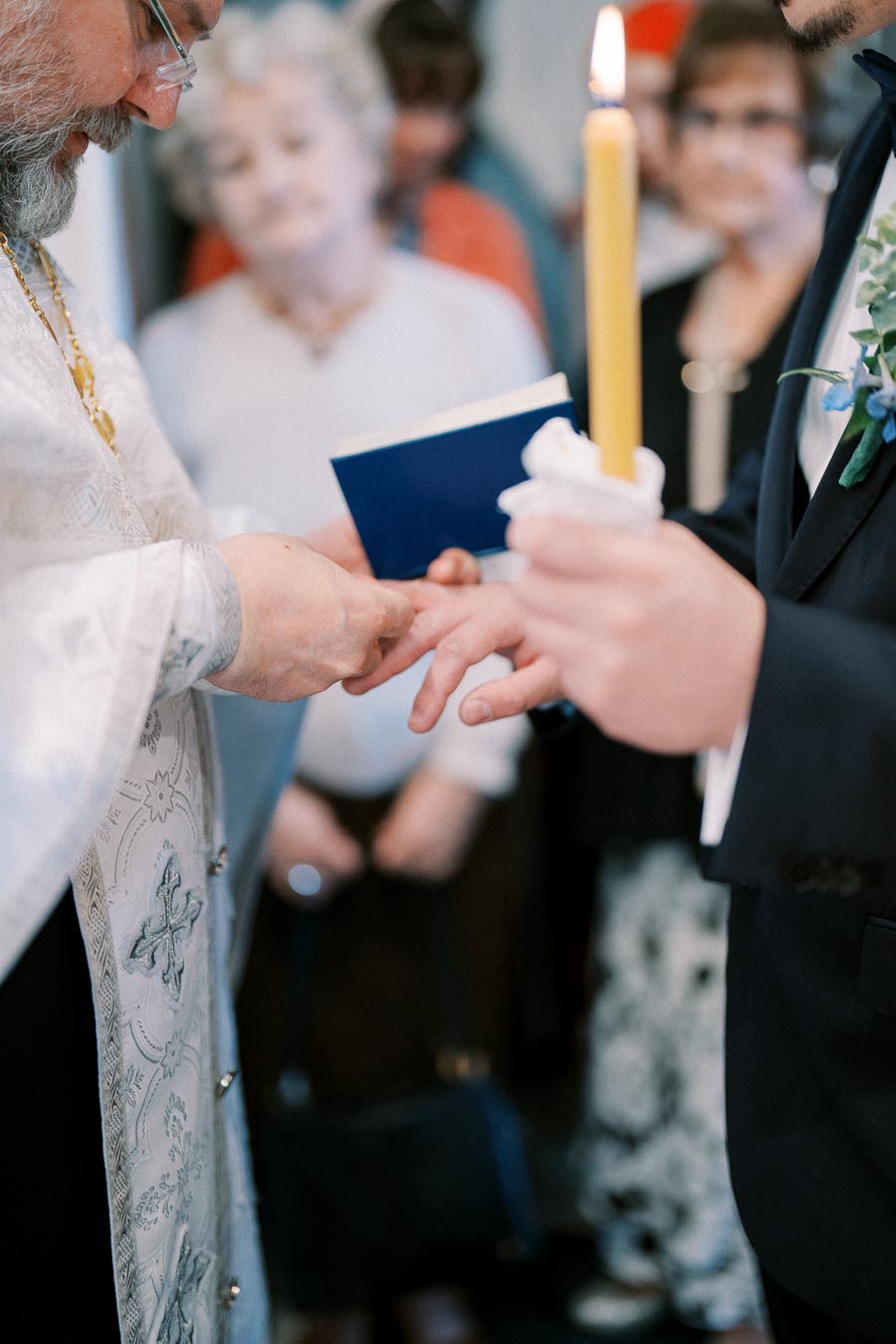 Priest placing a ring on a groom's finger during a wedding ceremony, with the priest holding a religious book and the groom holding a lit candle.