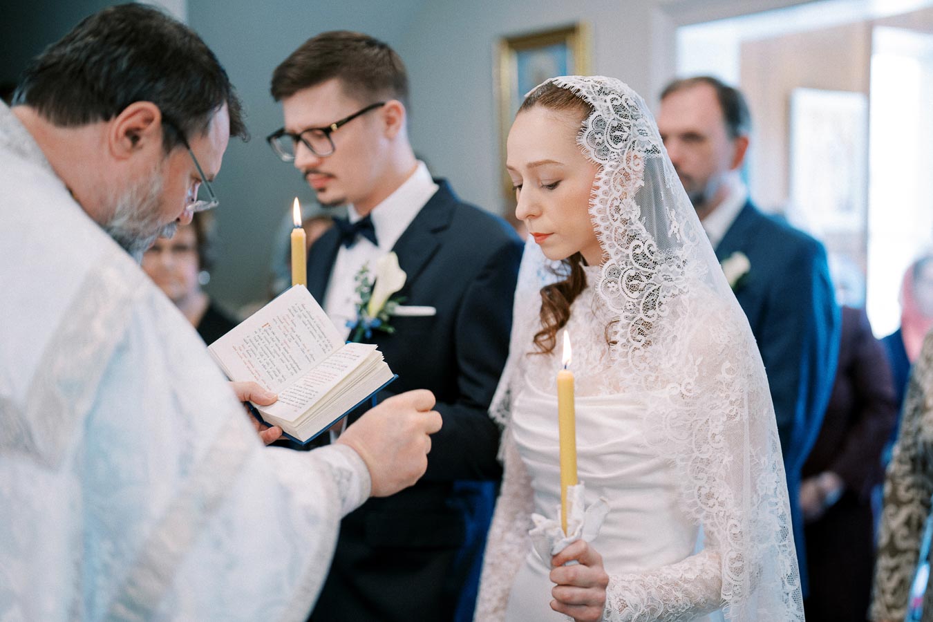 A bride and groom in traditional wedding attire hold candles during a ceremony, with a priest reading from a book. The bride wears a lace veil, emphasizing a serene and solemn atmosphere.