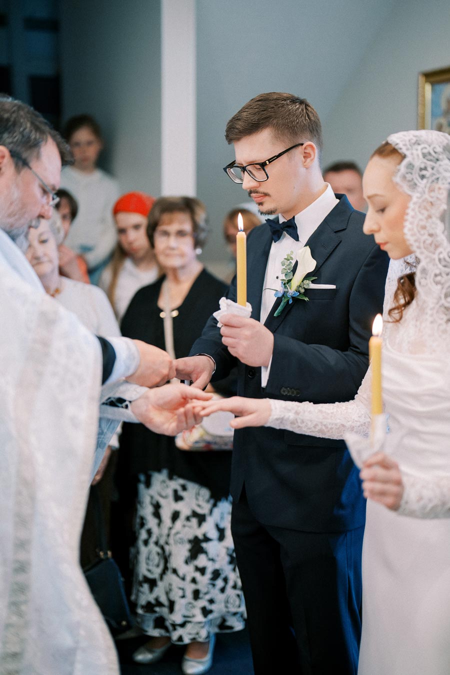 A bride and groom holding candles participate in a traditional wedding ceremony surrounded by family and friends in a church setting.