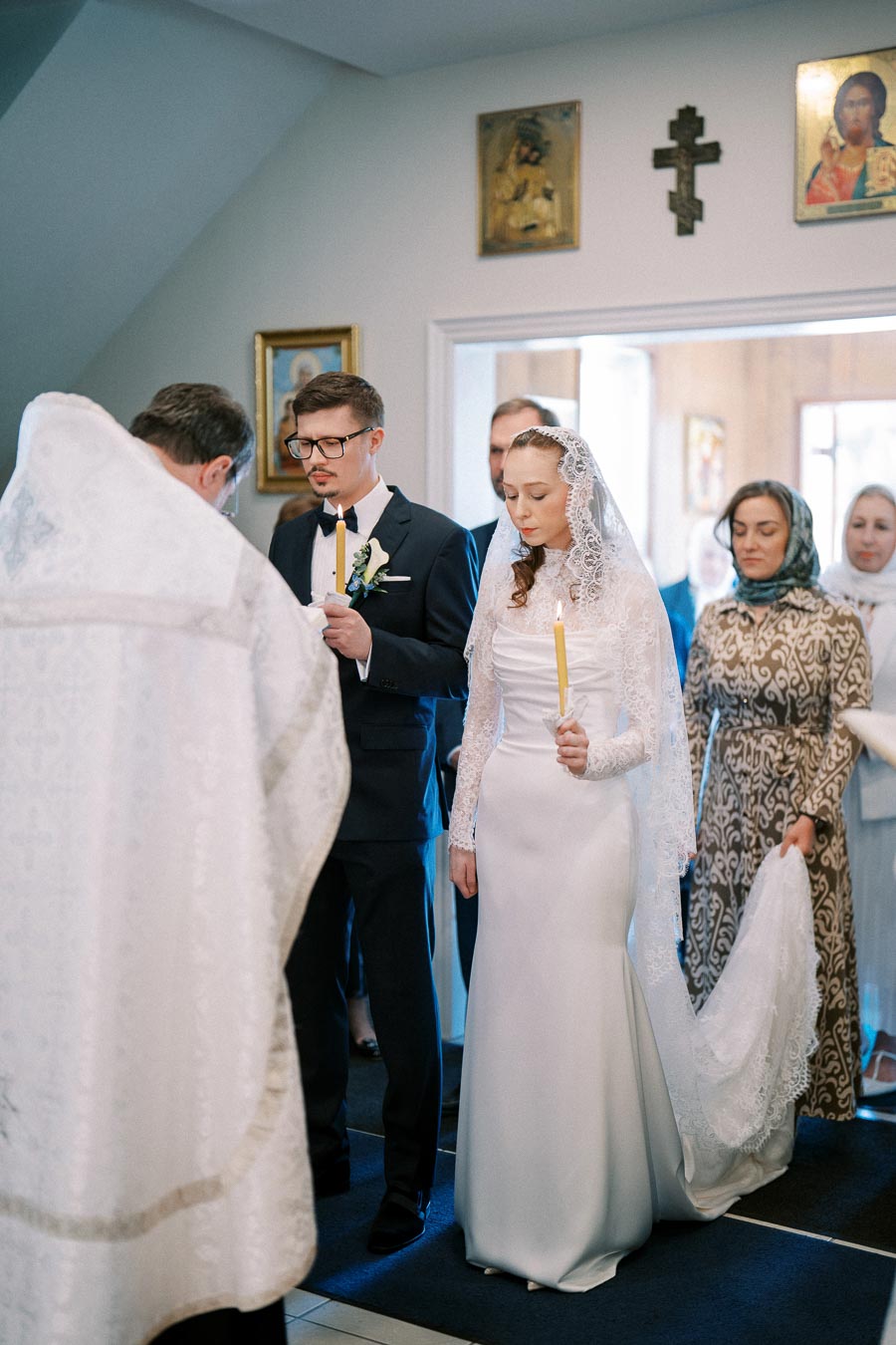 Orthodox wedding ceremony with bride and groom holding candles, dressed traditionally, surrounded by attendees in a decorated church setting.