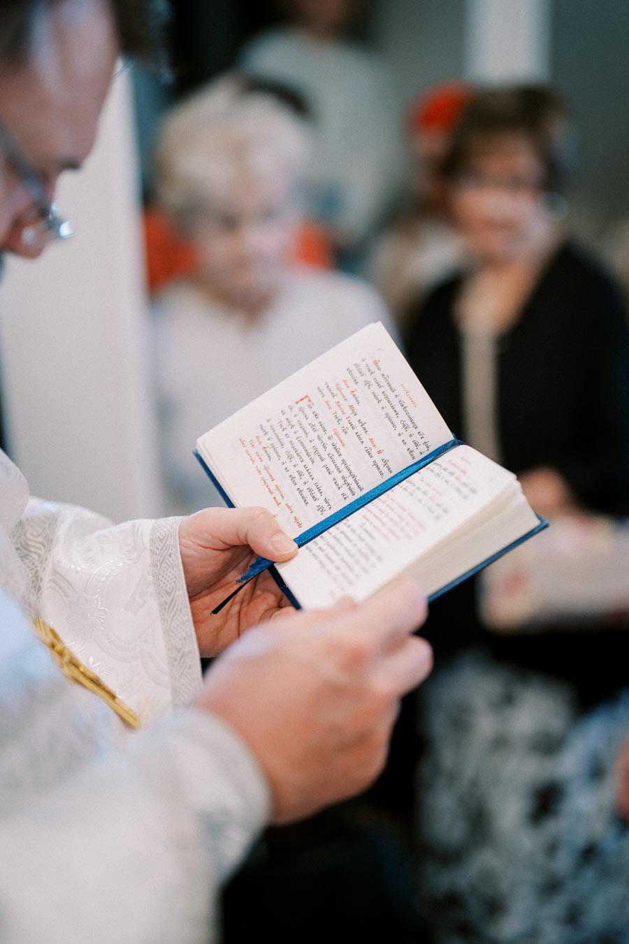 Priest reading from a book during a religious ceremony, with blurred people in the background.