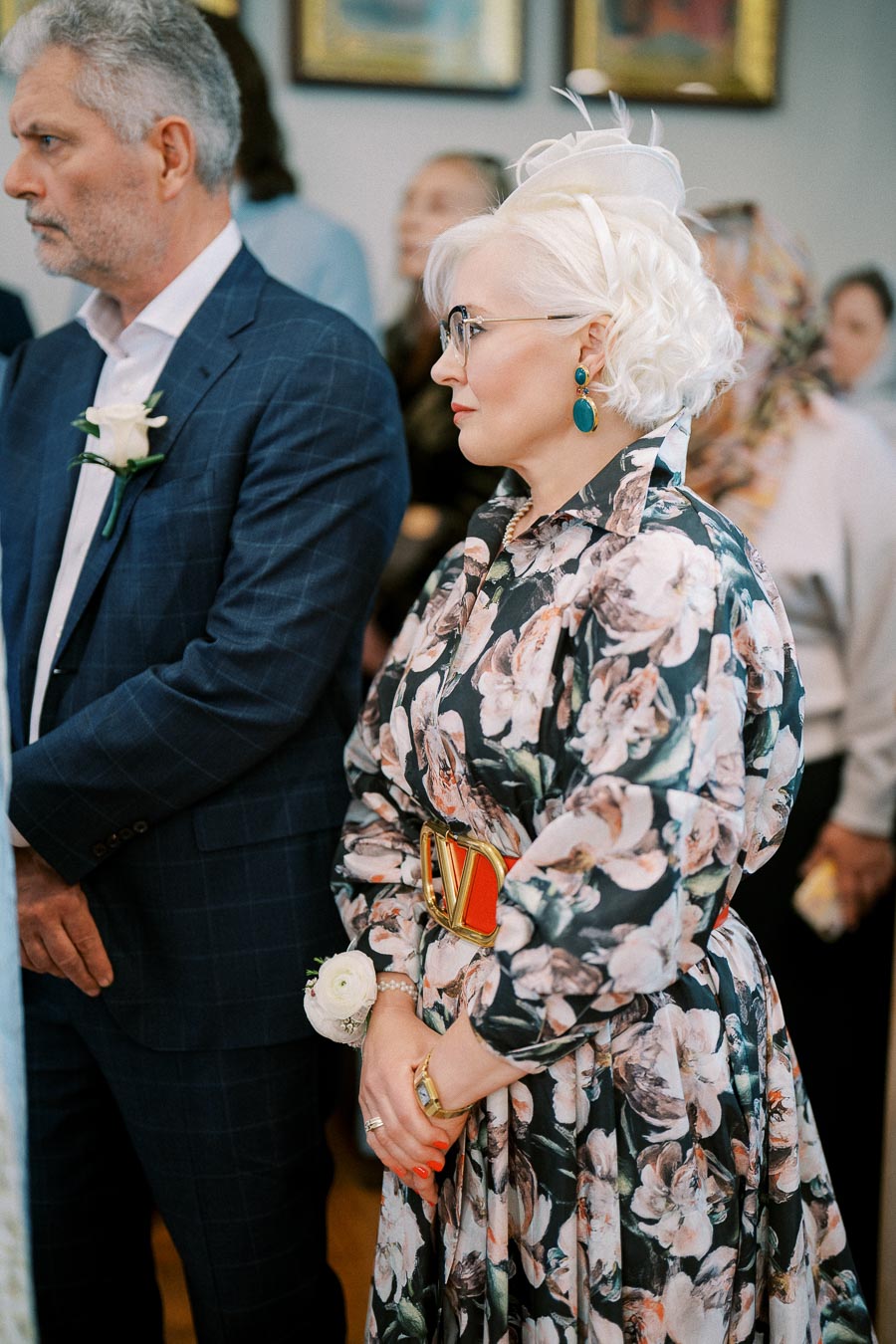 Elegant woman in floral dress and hat standing at formal event beside man in suit.