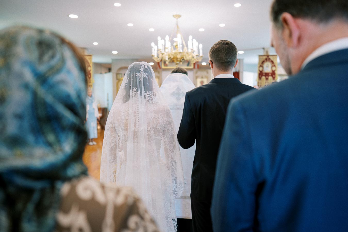 Back view of a bride and groom in formal attire walking towards an altar during a church wedding ceremony, with guests in attendance under a decorative chandelier.