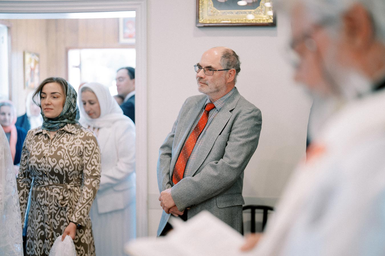 A group of people attending a formal indoor gathering, with a man in a gray suit and red tie standing attentively, and a woman in a patterned outfit with a headscarf holding a cloth.